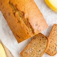 Overhead view of slices of banana bread cut from a loaf on a parchment-lined cutting board, next to yellow bananas.