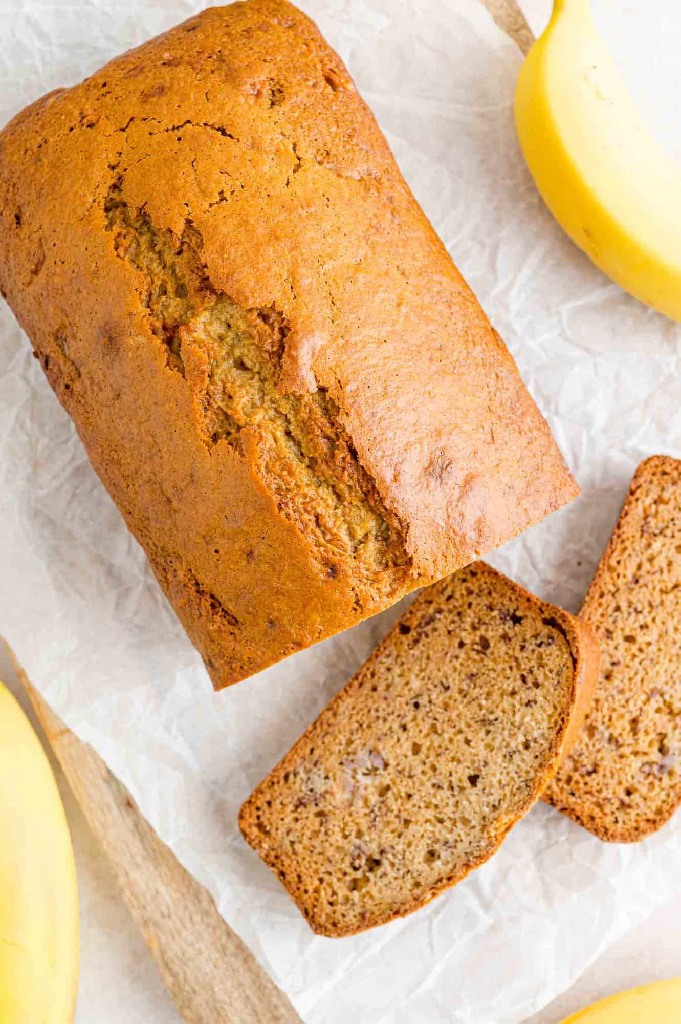 Overhead view of slices of banana bread cut from a loaf on a parchment-lined cutting board, next to yellow bananas.