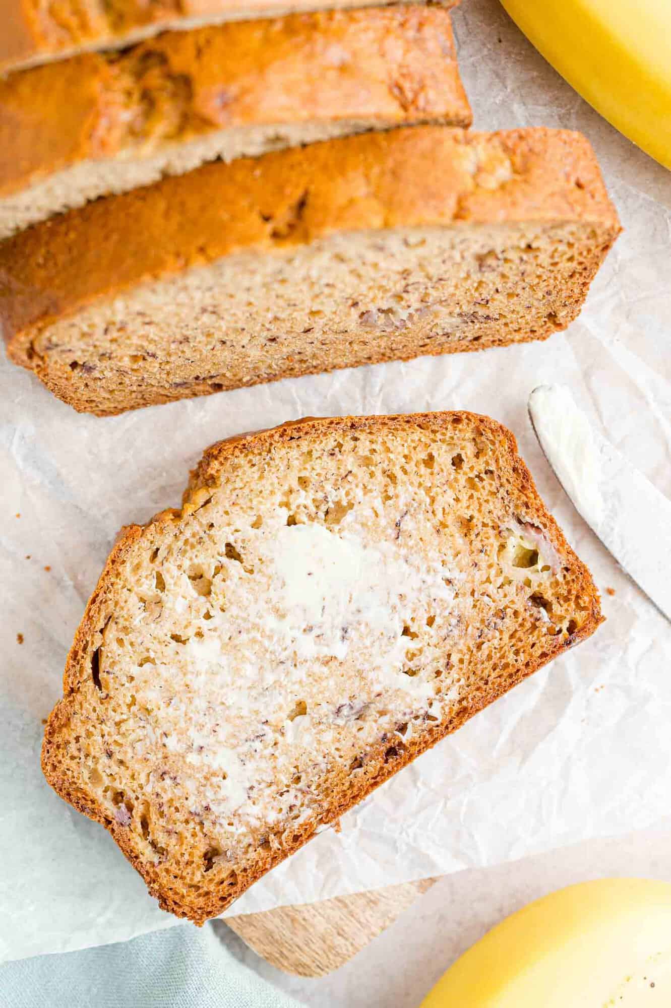 Overhead view of a slice of buttered banana bread laying next to a loaf on a parchment-lined cutting board.