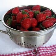Metal colander containing fresh strawberries on red checked cloth.