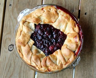 Overhead view of a rustic berry pie, crust pulled up around edges, berries showing in the middle, on a wooden background.