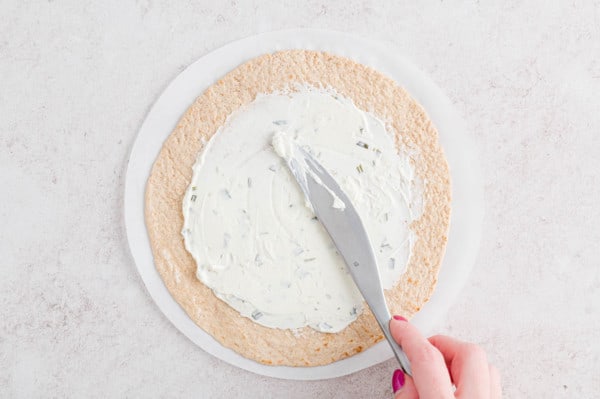 A hand using a knife to spread onion and chive cream cheese over a flour tortilla.