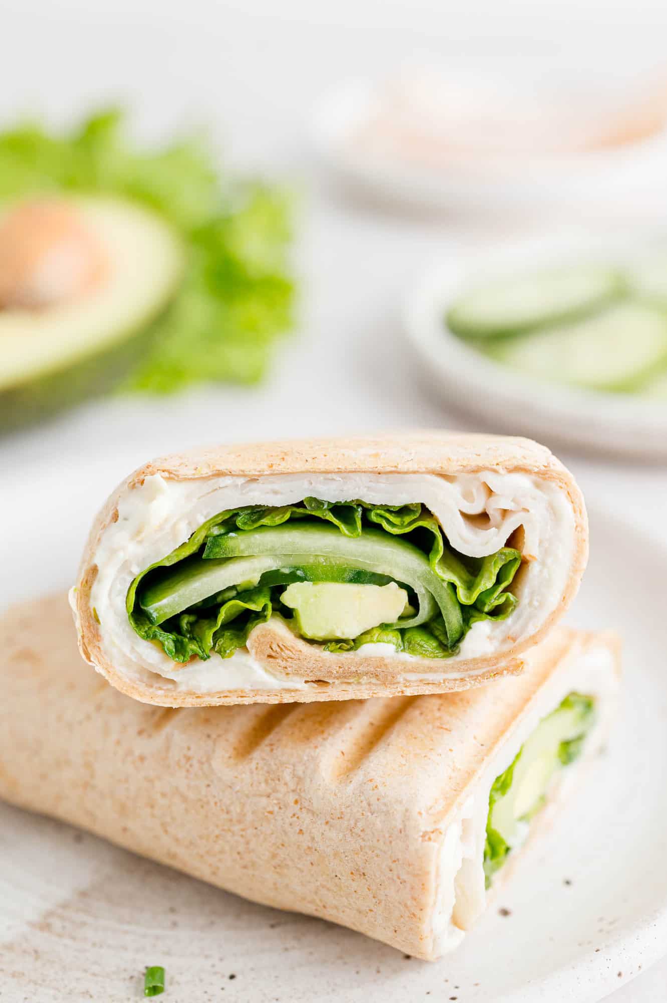 Two halves of a turkey wrap on a white plate, with an avocado and bowl of cucumber slices in the background.