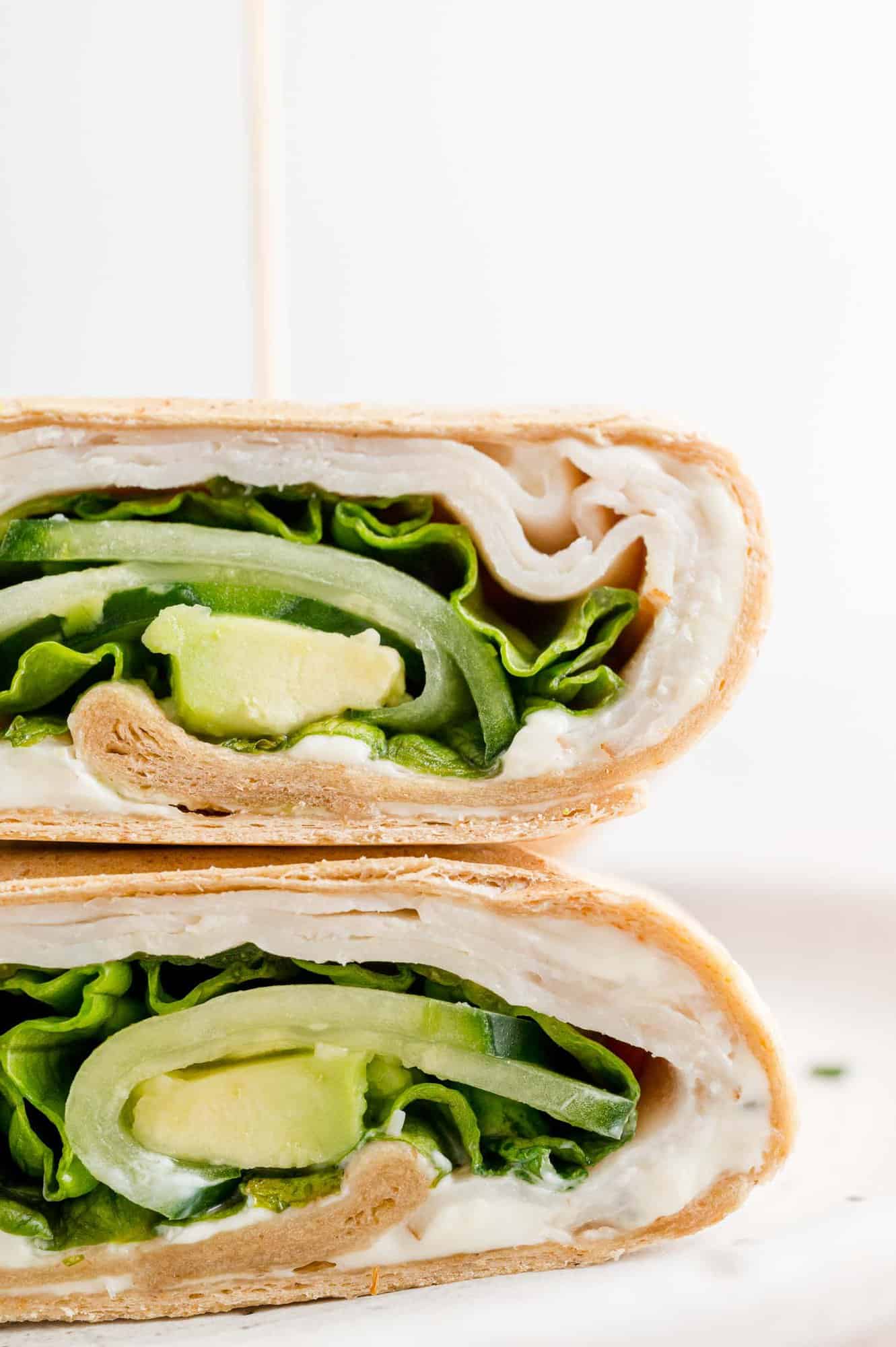 Make your own turkey wrap sandwich with spreadable cream cheese, deli-sliced turkey, lettuce, cucumbers, and avocado. Perfect for lunches! Close up of two halves of a turkey wrap stacked on a white plate.