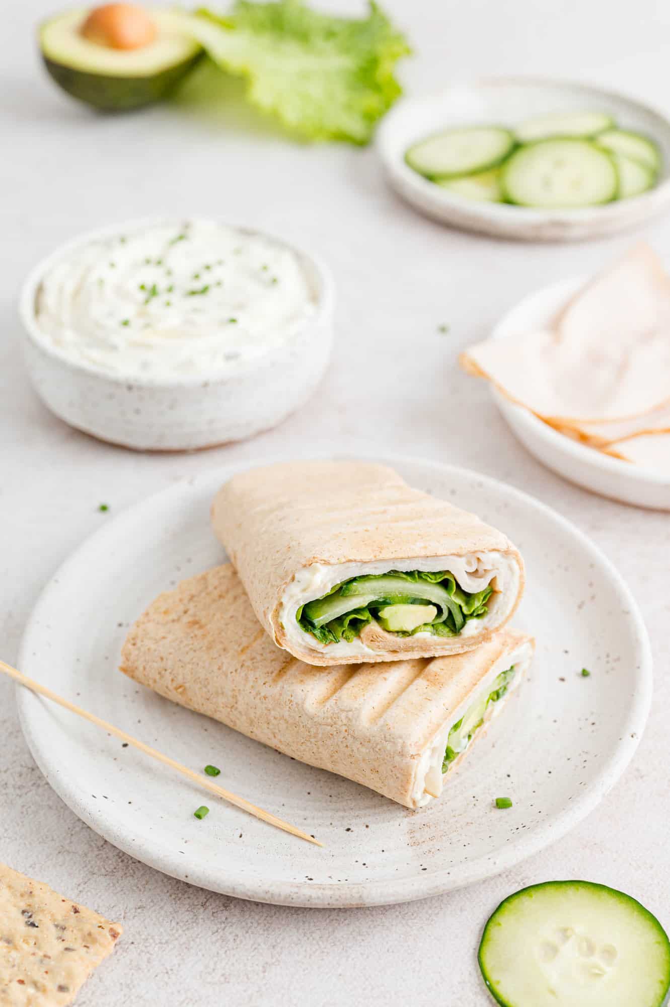 Two halves of a turkey wrap on a white plate, surrounded by bowls of ingredients.