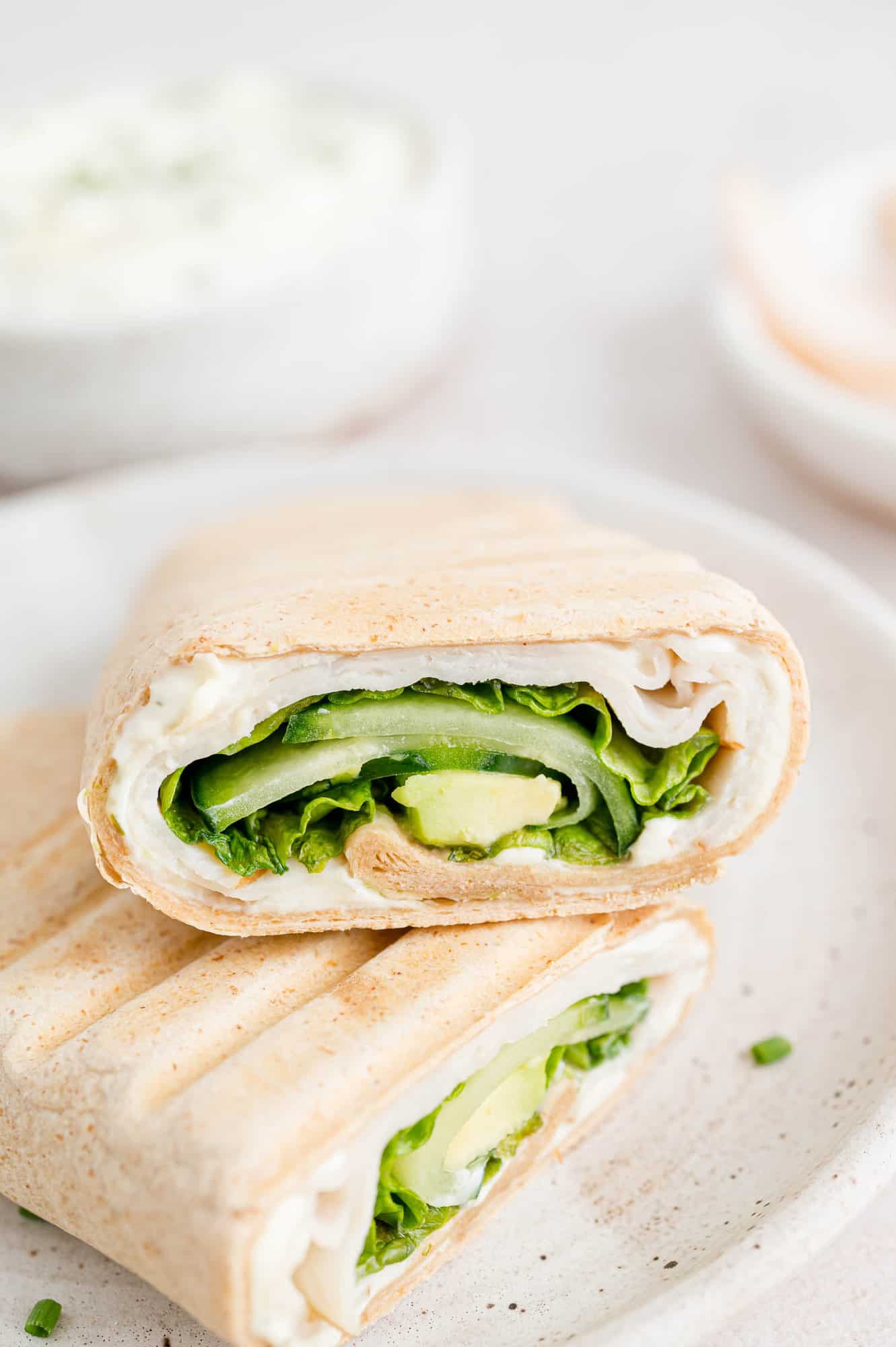 Make your own turkey wrap sandwich with spreadable cream cheese, deli-sliced turkey, lettuce, cucumbers, and avocado. Perfect for lunches! Two halves of a turkey wrap on a white plate.
