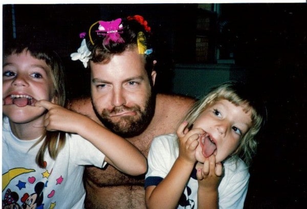 A dad with clips in his hair, next to his two daughters, making silly faces. 