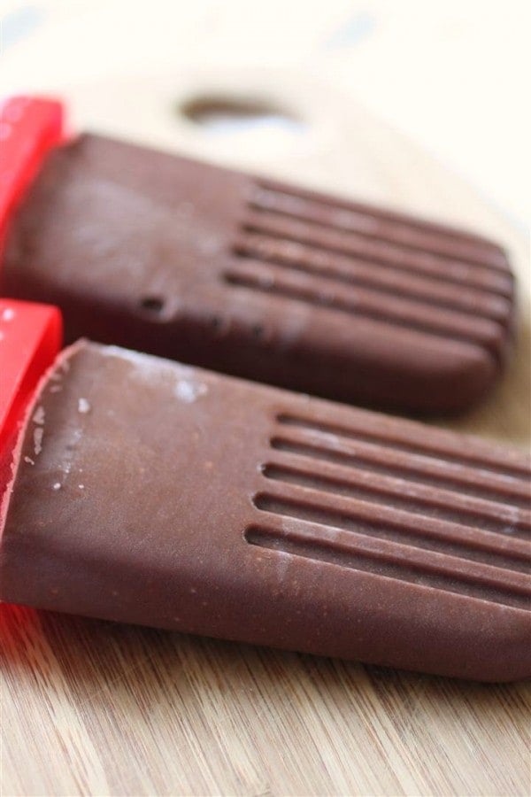 Side view close up of two chocolate pudding pops with red handles on bamboo cutting board.