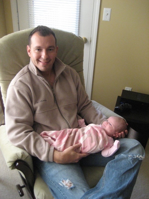 A young dad sitting in a green rocking chair, holding a baby dressed in all pink.
