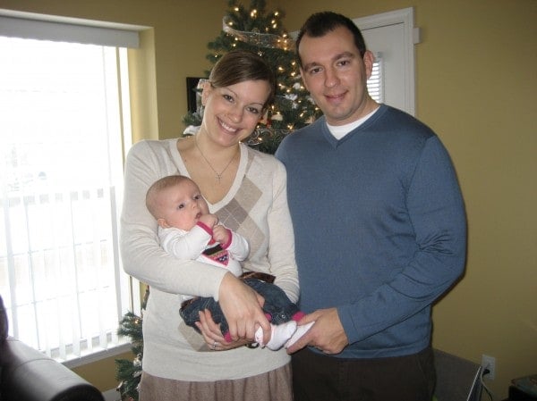 A young couple, standing and holding their baby in front of a Christmas tree.