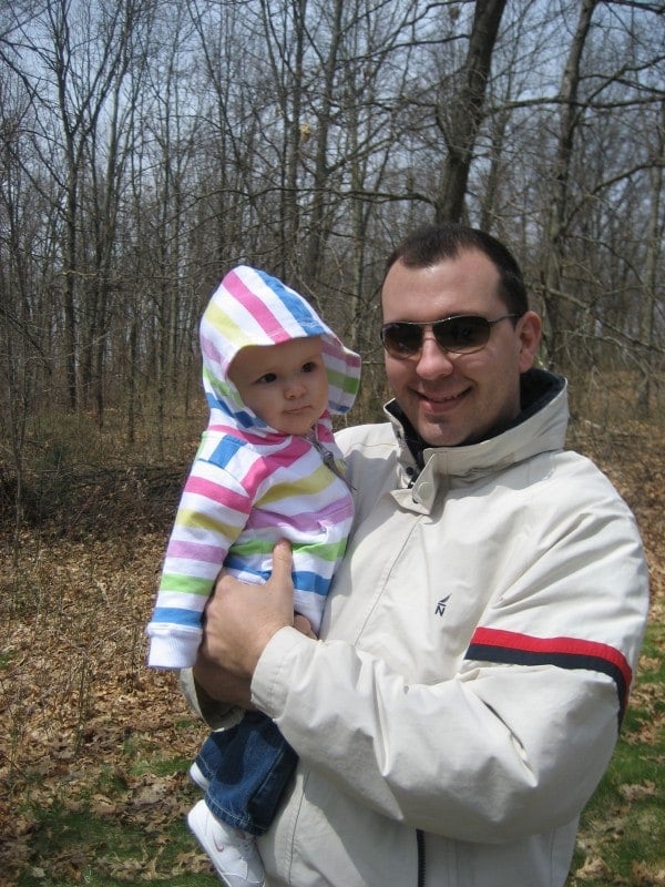 A man standing outside, holding a young baby girl in a striped hooded sweatshirt.