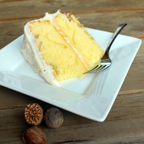 Front view of a slice of layer cake on square white plate with fork. In background is wooden deck boards and nutmegs.