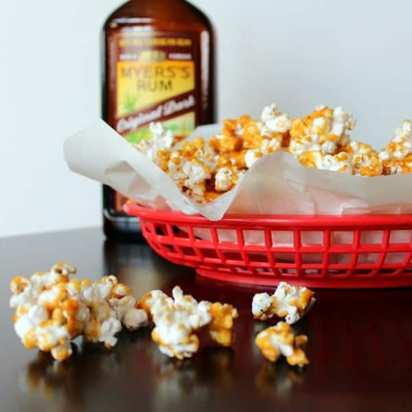 Caramel corn in paper lined red basket, with some on table in front of bowl, A bottle of Myers's rum in background.