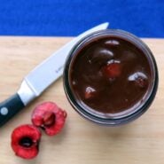 Overhead view of jar containing Black Forest hot fudge. In the background is a sharp knife, cutting board, and one cherry cut in half.