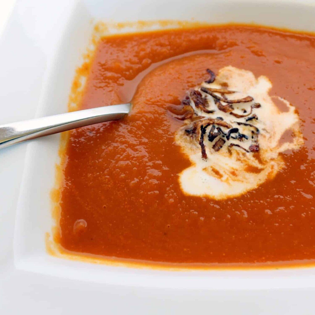 Square white bowl containing carrot soup with a swirl of sour cream and sprinkled with crispy shallots.