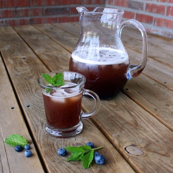 Front view of clear glass and pitcher containing blueberry iced tea, garnished with mint sprig, on wooden deck boards.