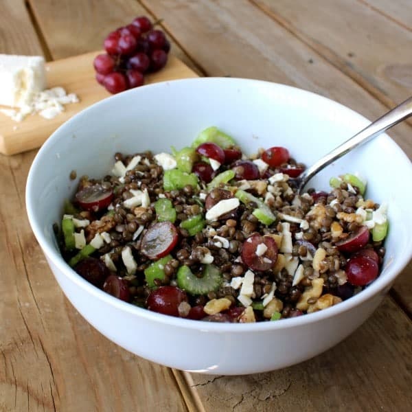 A large white bowl full of a lentil salad with lentils, celery, feta, grapes, and walnuts. Grapes and feta are also pictured in the background.
