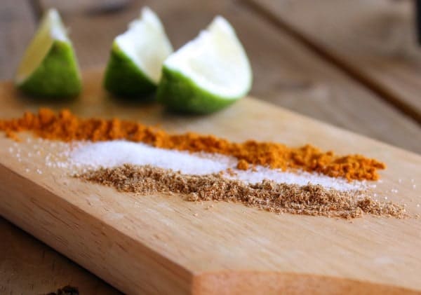 Closeup horizontal shot of wooden cutting board with three rows of spices, and 3 lime wedges.