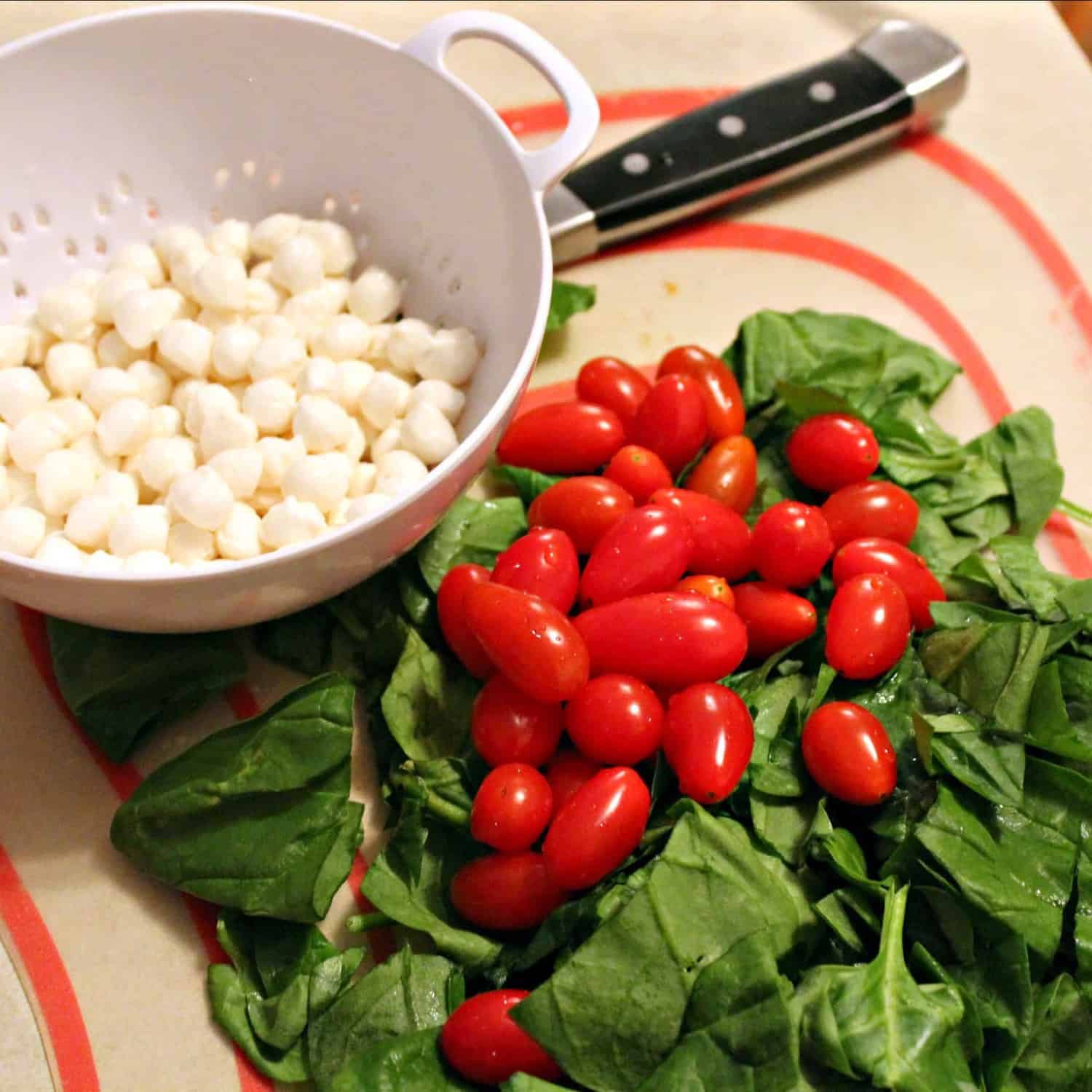 Overhead shot of a pile of fresh spinach leaves, with several grape tomatoes piled on top. Along side is a small white colander containing many mozzarella pearls.