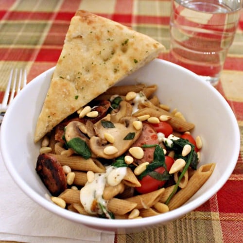 White bowl with pasta, a wedge of bread balanced on edge, on plaid tablecloth with water glass, fork, and napkin in background