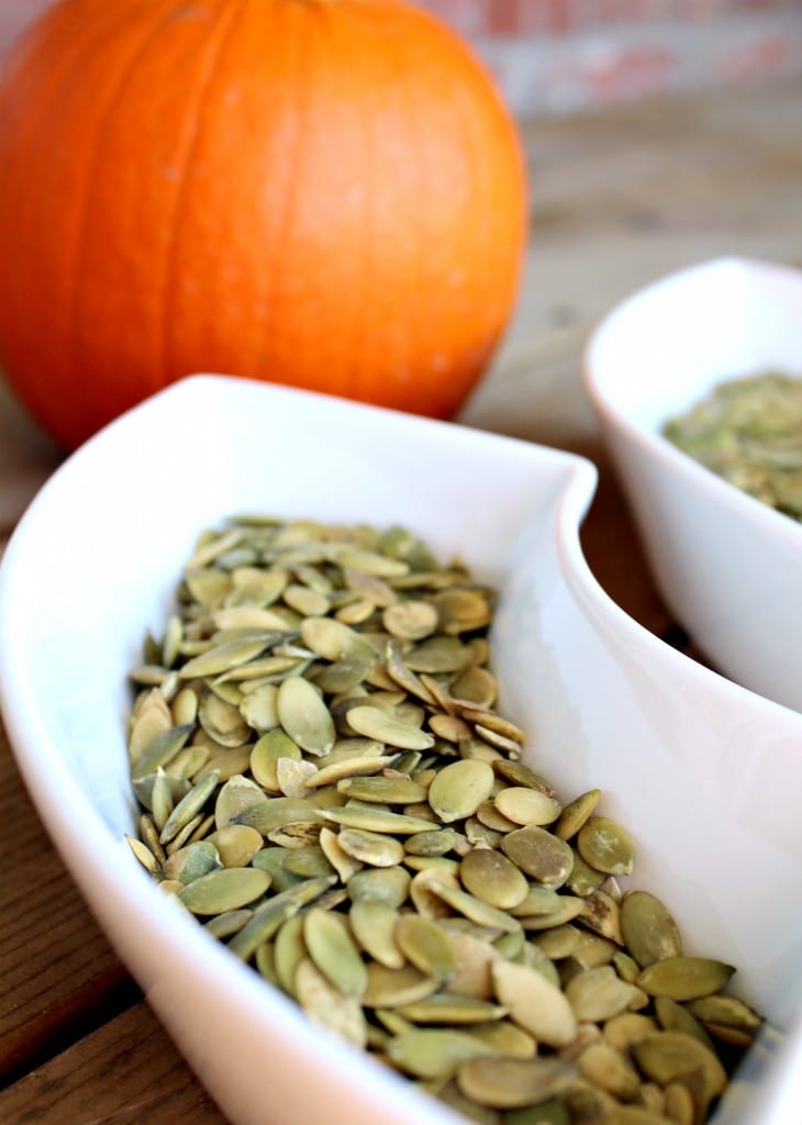 Curved white dish containing untoasted pepitas. Pumpkin in background.