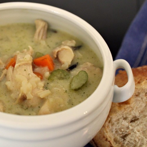 White crock-style bowl with creamy soup in it. Bits of chicken, rice, celery, and carrots are visible.