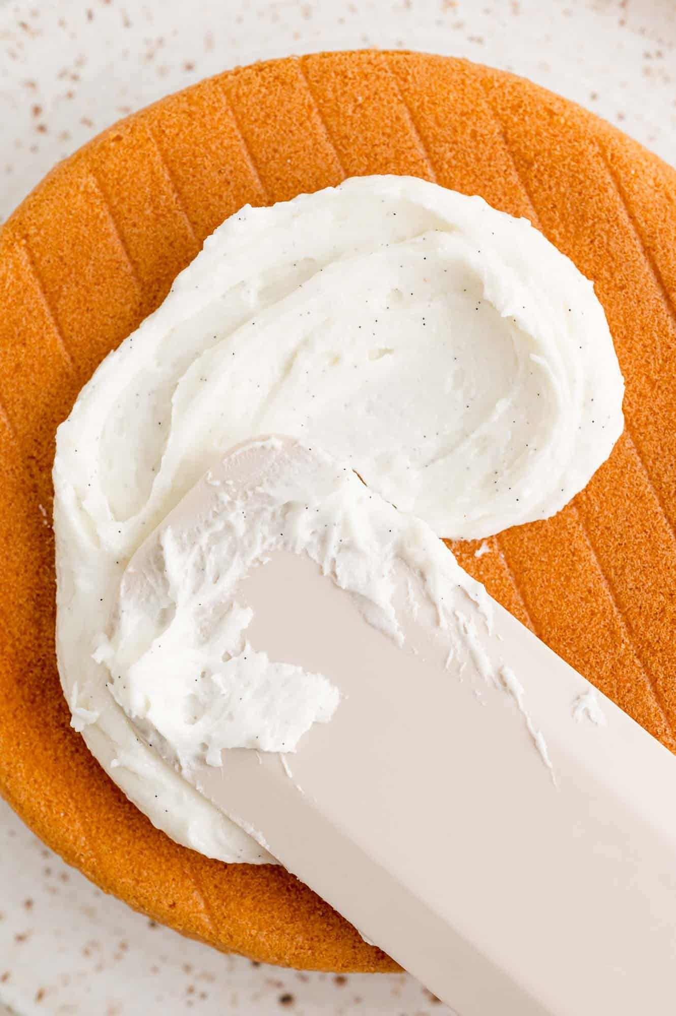 A spatula spreads vanilla buttercream over a vanilla cake layer.