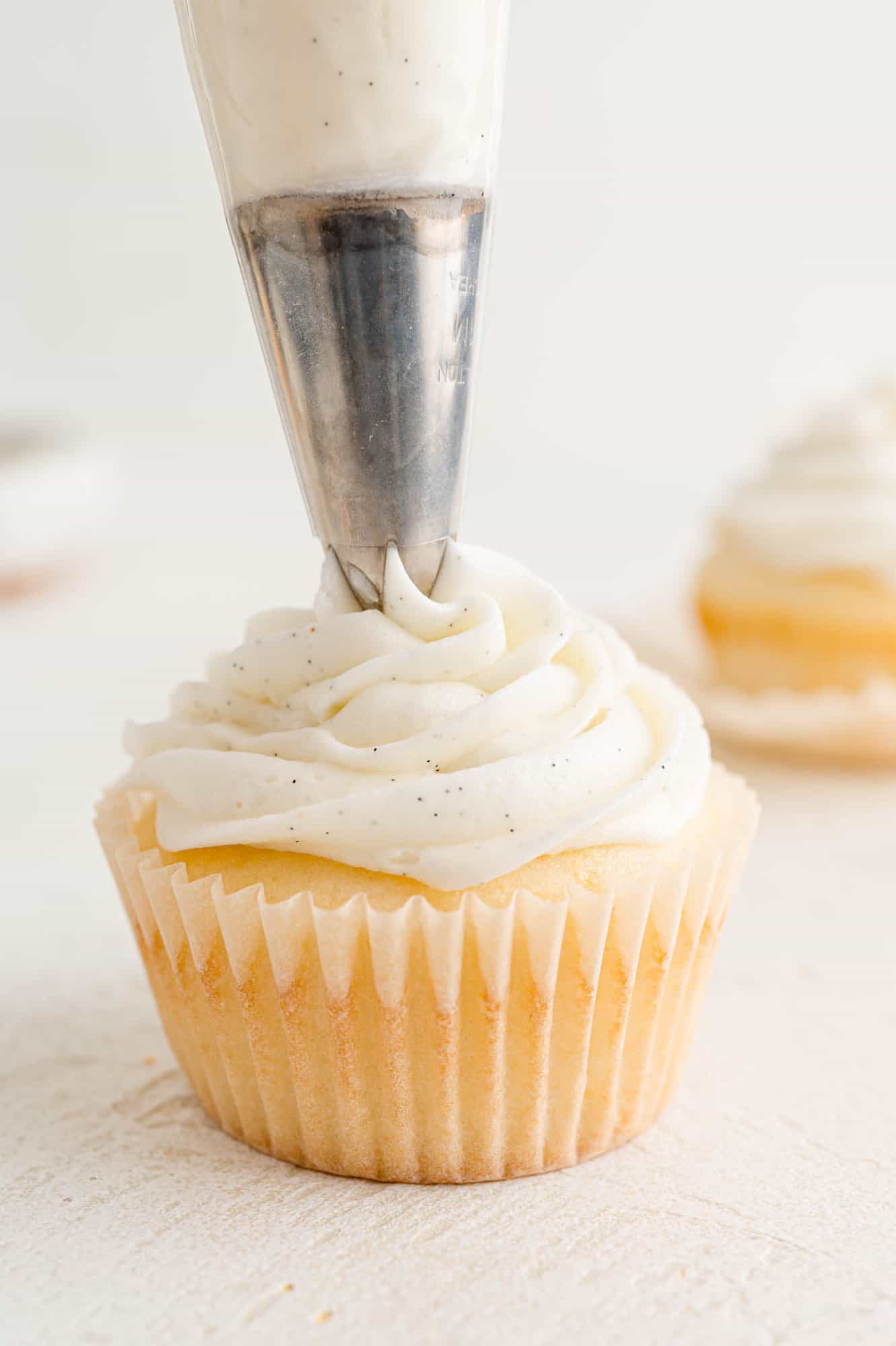 A piping tip pipes a swirl of vanilla buttercream onto a vanilla cupcake.