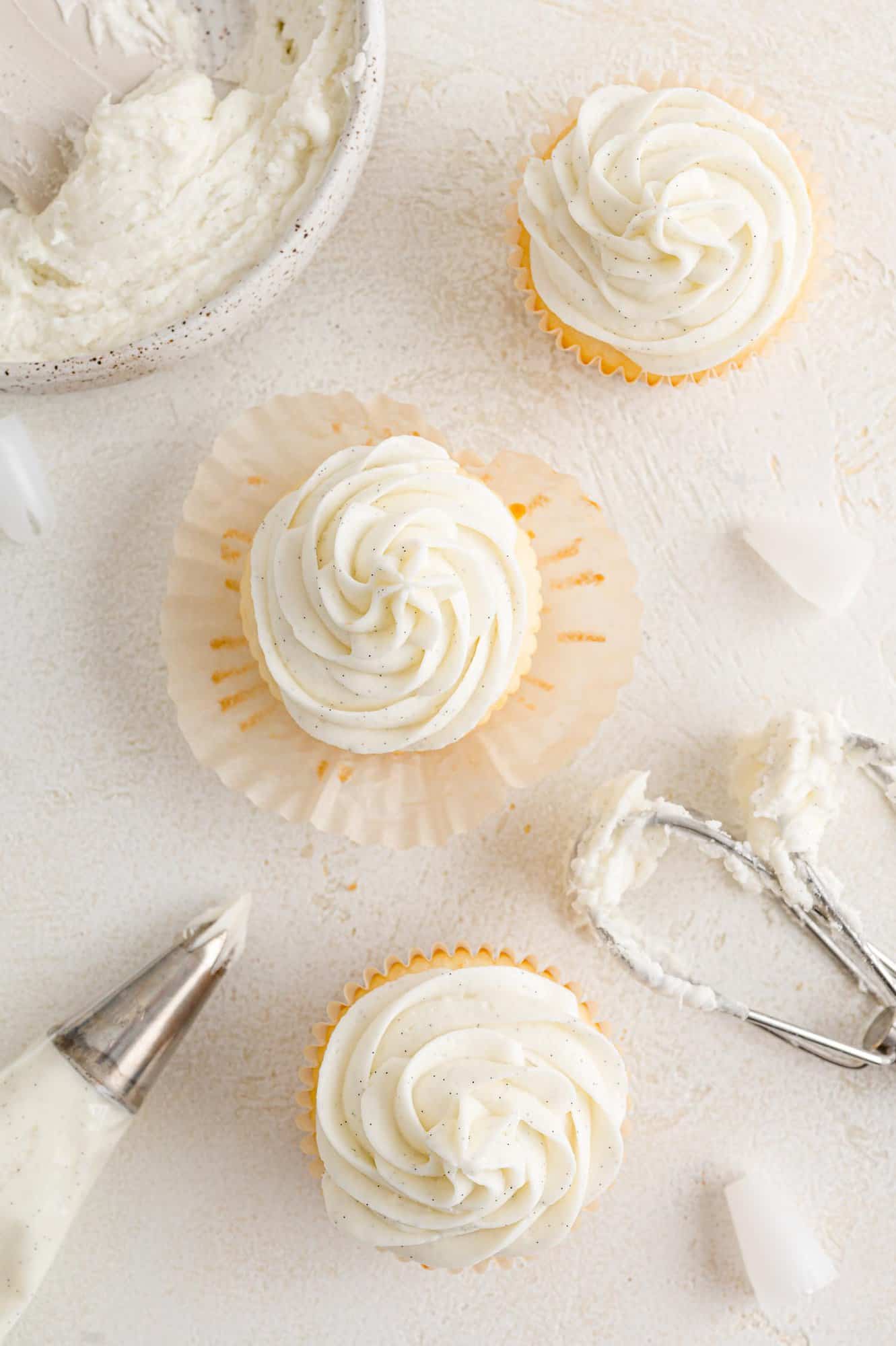 Overhead view of vanilla cupcakes topped with swirls of vanilla buttercream frosting, next to a piping bag and a mixer attachment.