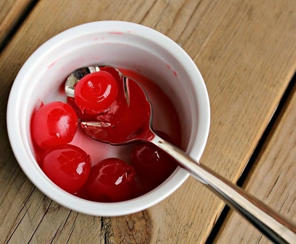Overhead view of small white bowl containing a spoon and 5 maraschino cherries.