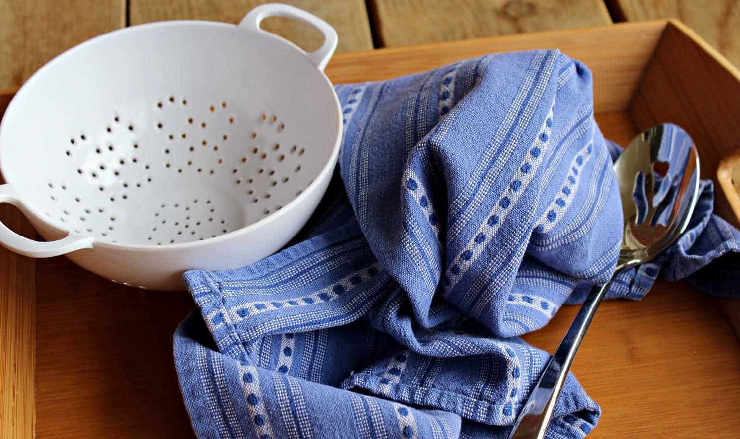 White strainer, blue towel, and slotted spoon on a wooden tray.
