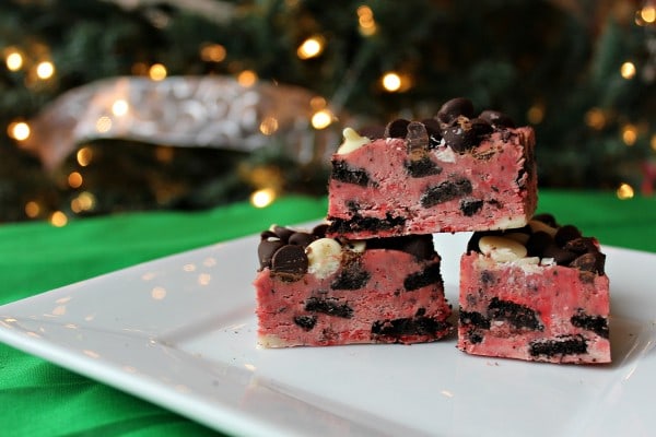 Three pieces of red fudge flecked with cookies and cream pieces, as well as chocolate chips and white chocolate chips. A holiday tree is visible in the background.
