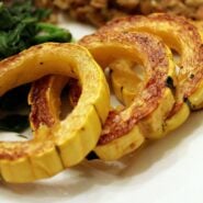 Four rings of roasted delicata squash on a white plate, with other food visible in background.