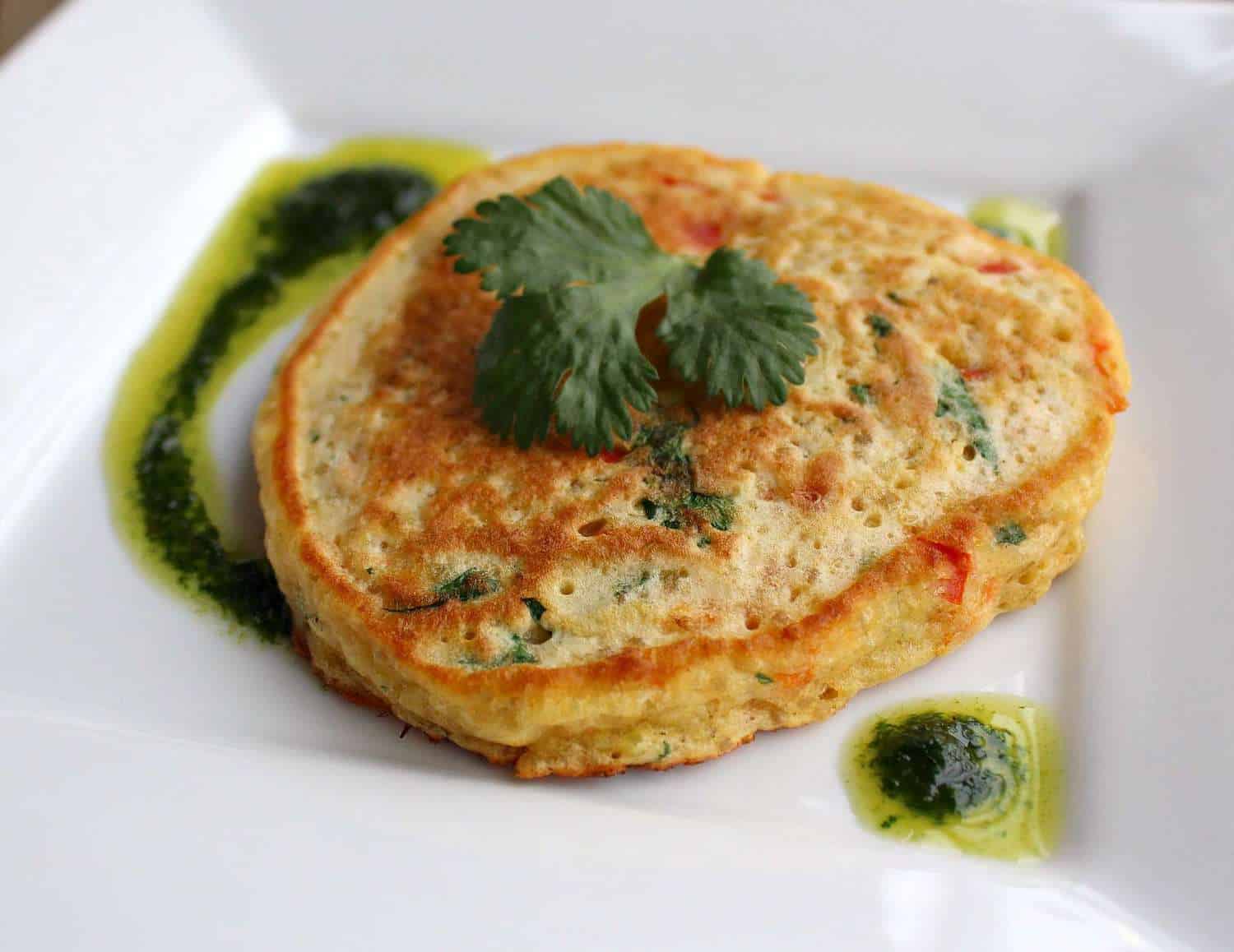 A single pancake on square white plate with vinaigrette, garnished with sprig of parsley.