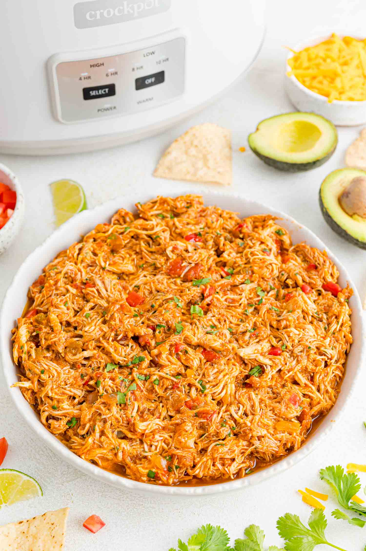 A large bowl of crockpot salsa chicken on a countertop next to the crockpot, surrounded by garnishes.