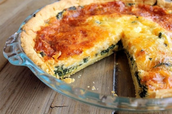 Image of a spinach quiche in a glass pan with one slice removed. 