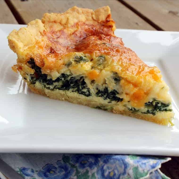 Close up image of a slice of quiche on a white plate. Quiche has a cornmeal crust and is filled with eggs, swiss cheese, and spinach.