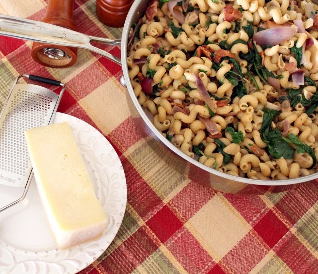 Overhead view of large skillet with pasta dish, small white plate containing Parmesan cheese and grater, on a red plaid tablecloth.