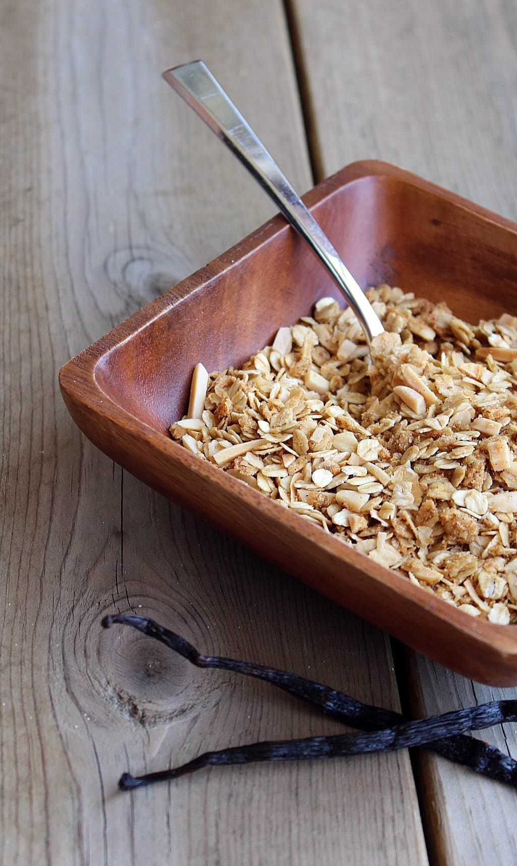 vanilla-bean-granola Partial view of square wooden bowl containing granola with spoon. In front of bowl are two vanilla beans.