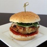 A turkey burger on a white plate, the burger has cheese on it, fresh basil, and pizza sauce.
