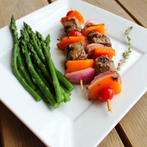 2 beef skewers on square white plate, with asparagus spears and sprig of thyme.