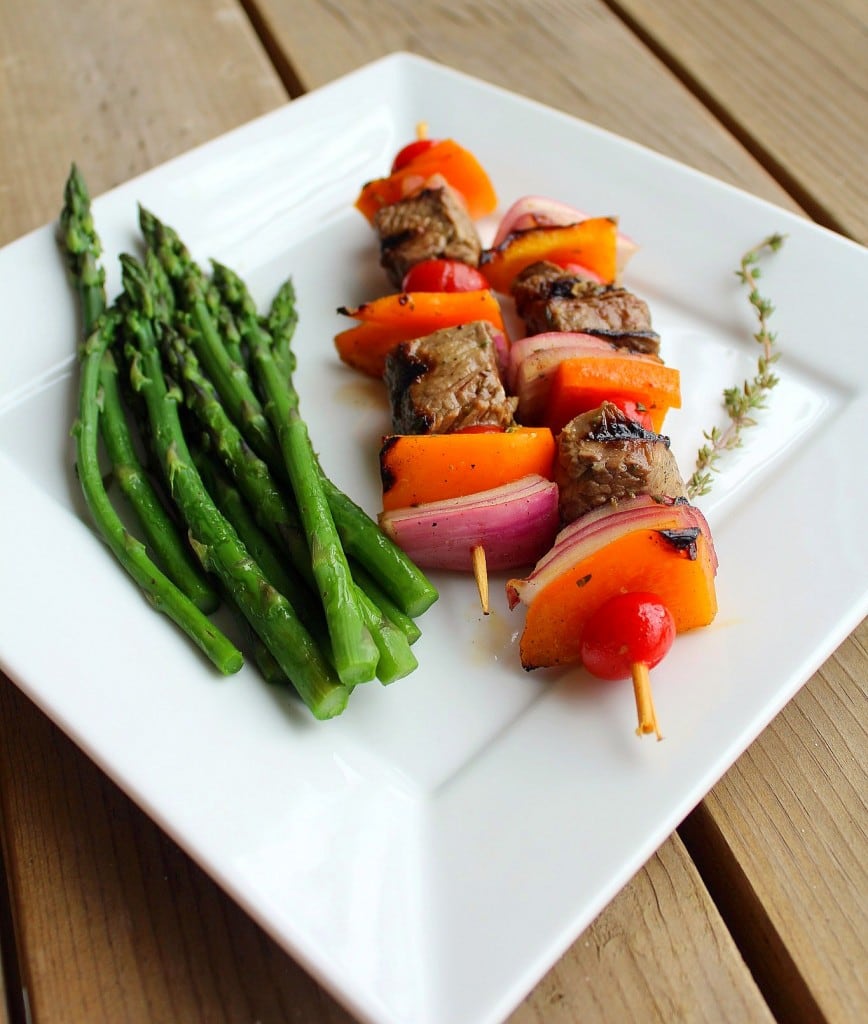 Two kabobs on square white plate, with asparagus spears and sprig of thyme.