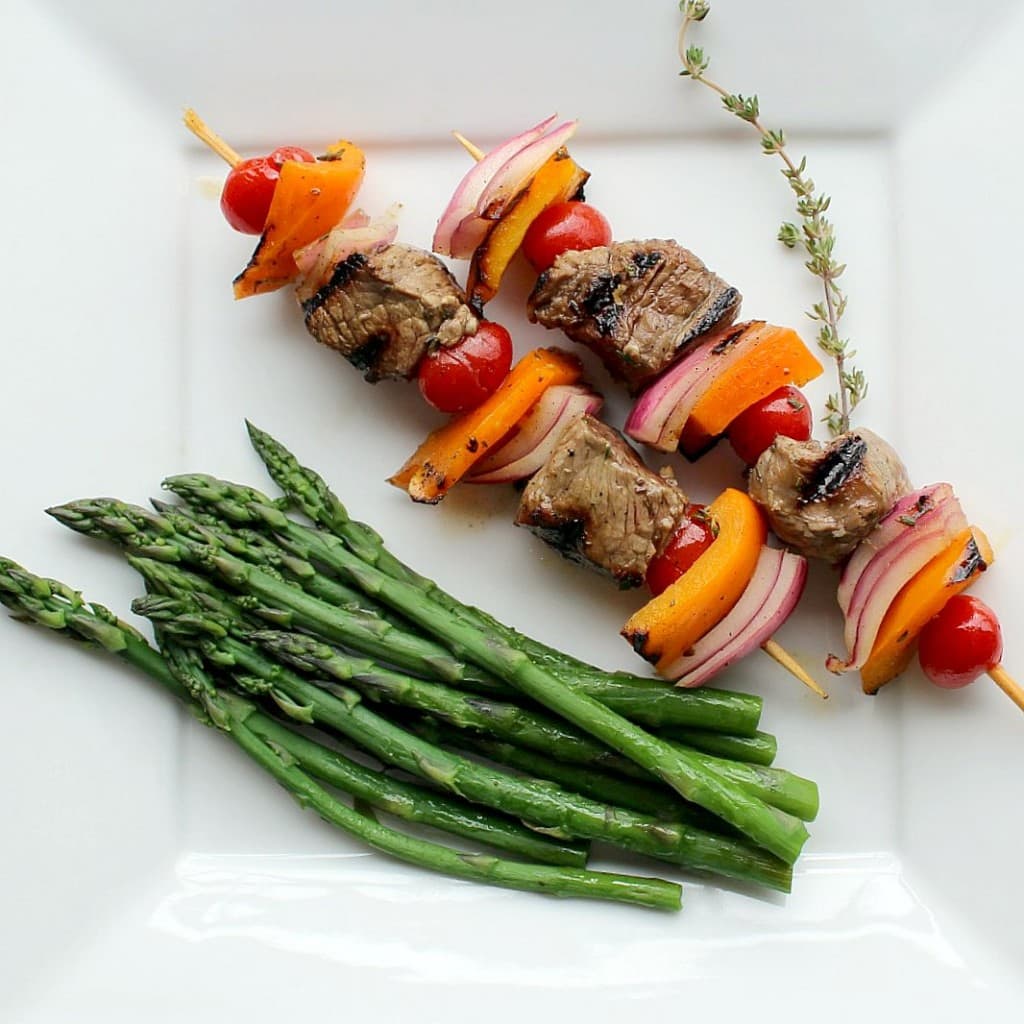 Two kabobs on square white plate with asparagus spears and sprig of thyme.