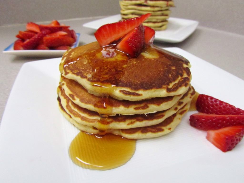 Stack of four pancakes with sliced berries and maple syrup, on white plate.