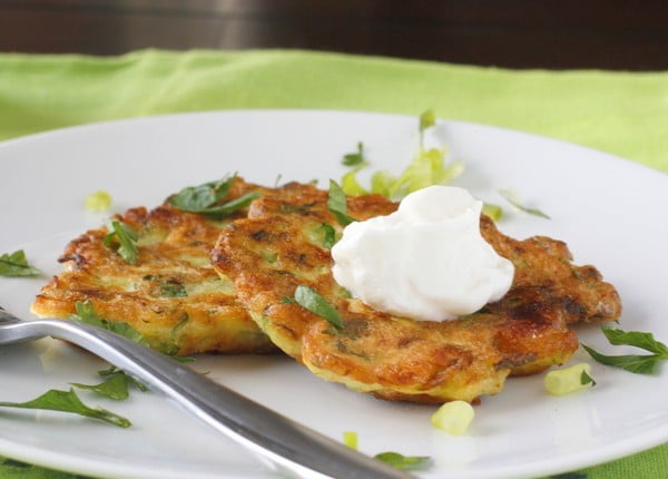 Two zucchini and feta pancakes on a white plate, garnished with sour cream and sliced scallions.