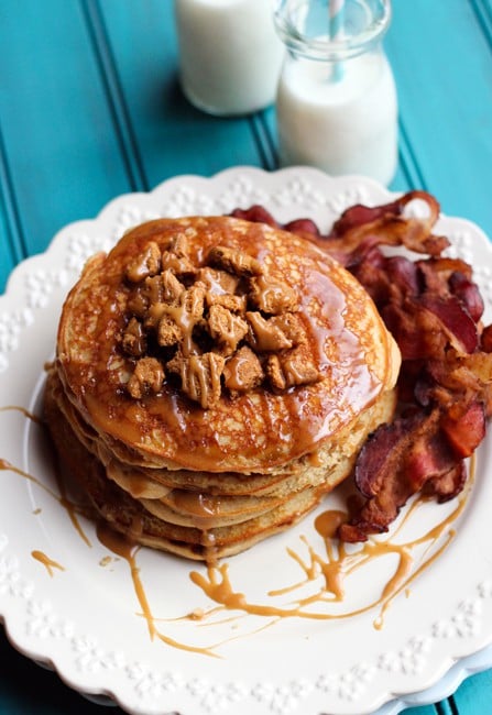 Stack of pancakes on white plate, with syrup and crushed cookies, and bacon on side, with glass of milk.
