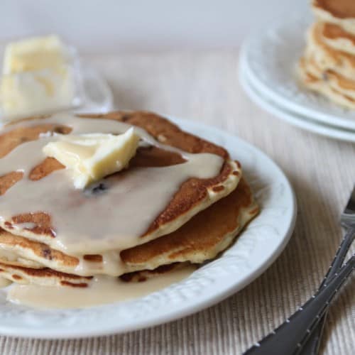 Stack of pancakes with butter and glaze on white plate with more pancakes in background.