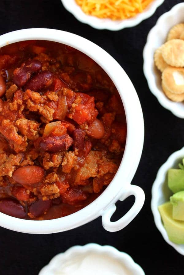 Image of white bowl of chili surrounded by small bowls of chili toppings, such as sour cream, diced avocado, oyster crackers, and shredded cheese.