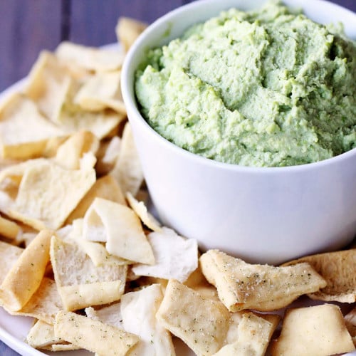 Closeup view of light green dip in a white bowl, surrounded by pita chips.