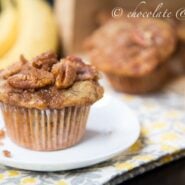 Front view of one banana streusel muffin on small white plate, with bananas and more muffins in background. Text overlay reads "chocolate & carrots".